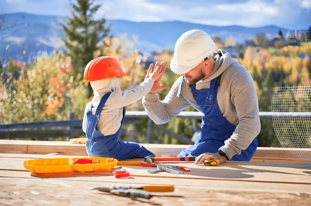 Gradbeni delavec v modrem kombinezonu in beli trdi kapi daje petko otroku v oranžni trdi kapi in modrem kombinezonu, obdanemu z orodjem na leseni ploščadi s slikovitim gorskim ozadjem pod jasnim modrim nebom.