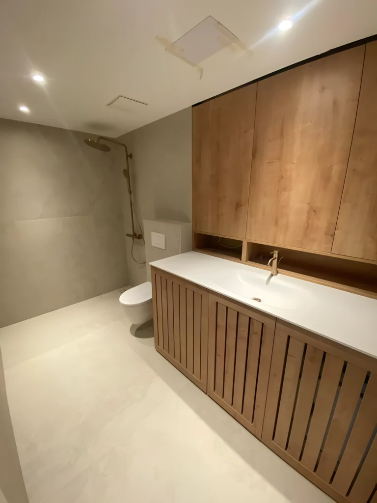 Elegantna sodobna kopalnica s stenami in tlemi, obloženimi s sivimi ploščicami, z belim straniščem, okroglim umivalnikom z zlato pipo na leseni toaleti in stropni tuš v prostoru za prhanje. Zasnova vključuje leseno omarico nad umivalnikom in vgradno razsvetljavo na stropu.