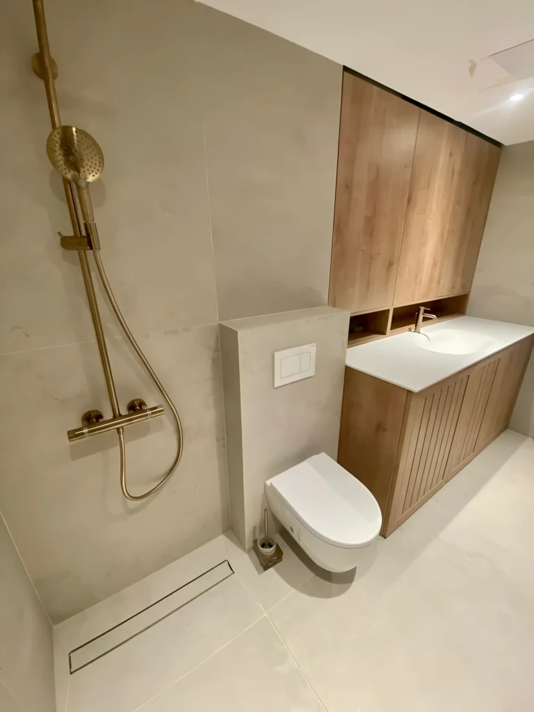 Moderna minimalistična kopalnica z zlatimi napeljavami, tušem, stenskim straniščem, leseno toaleto, belim pultom, ogledalno omarico ter stenami in tlemi, obloženimi z bež ploščicami. Vključuje dežno glavo za prhanje, linearni odtok in osvetlitev v toplih tonih za eleganten, sodoben dizajn.